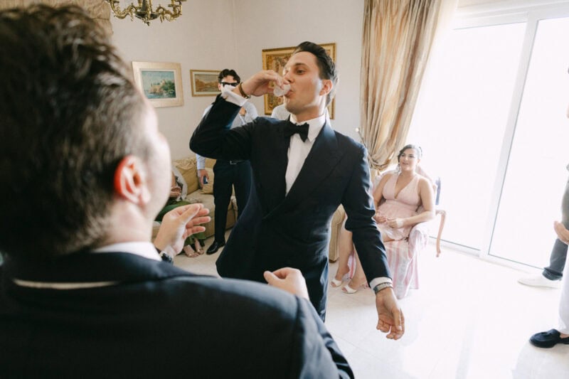 Groom in tuxedo drinking at wedding celebration indoors.