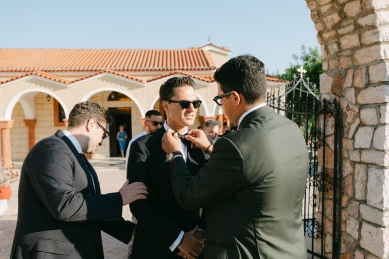 Groom and best Men in suits at outdoor event, engaging in conversation and adjusting ties before ceremony in messinian wedding