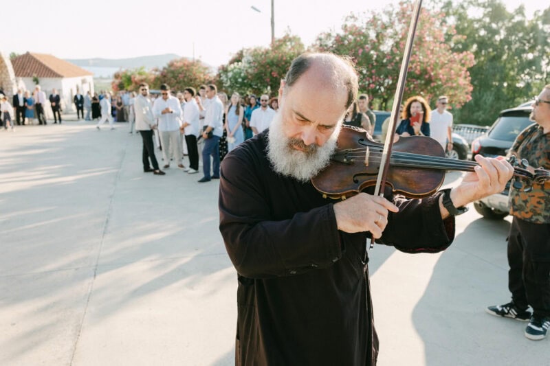 Priest performing violin outdoors before ceremony at a Messinian wedding with a group in the background.
